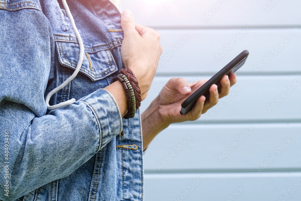 Unrecognizable woman holding smartphone in hand, texting messages, surfing internet and using social networks outdoors, she wear denim jacket and leather bracelets