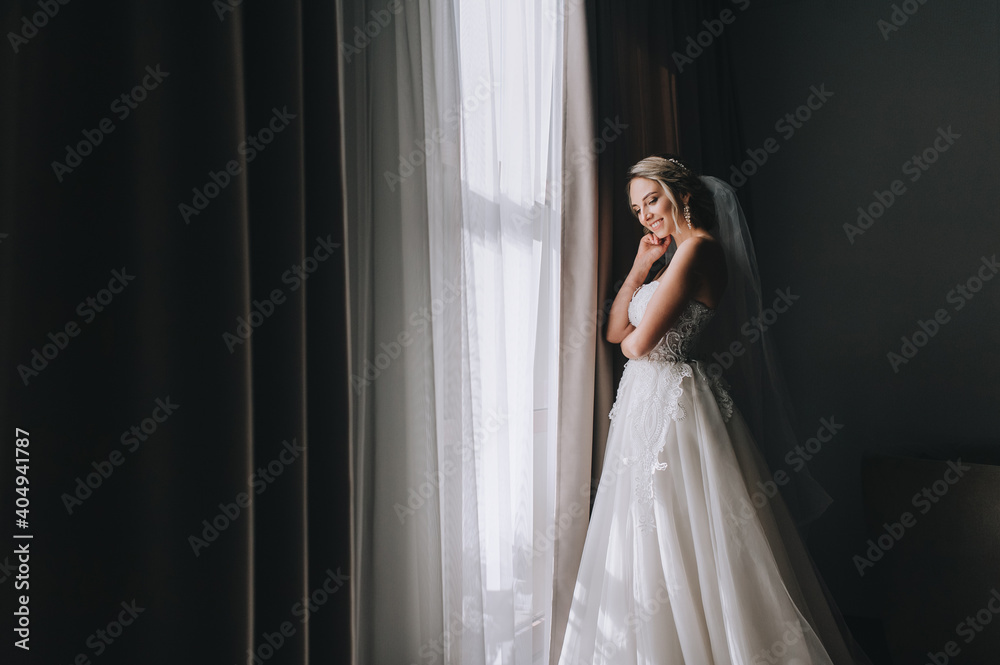 A beautiful blonde bride in a white long dress stands near the window with curtains in the room of a posh hotel. Wedding portrait. A beautiful blonde bride in a white long dress stands near the window with curtains in the room of a posh hotel. Wedding portrait.
