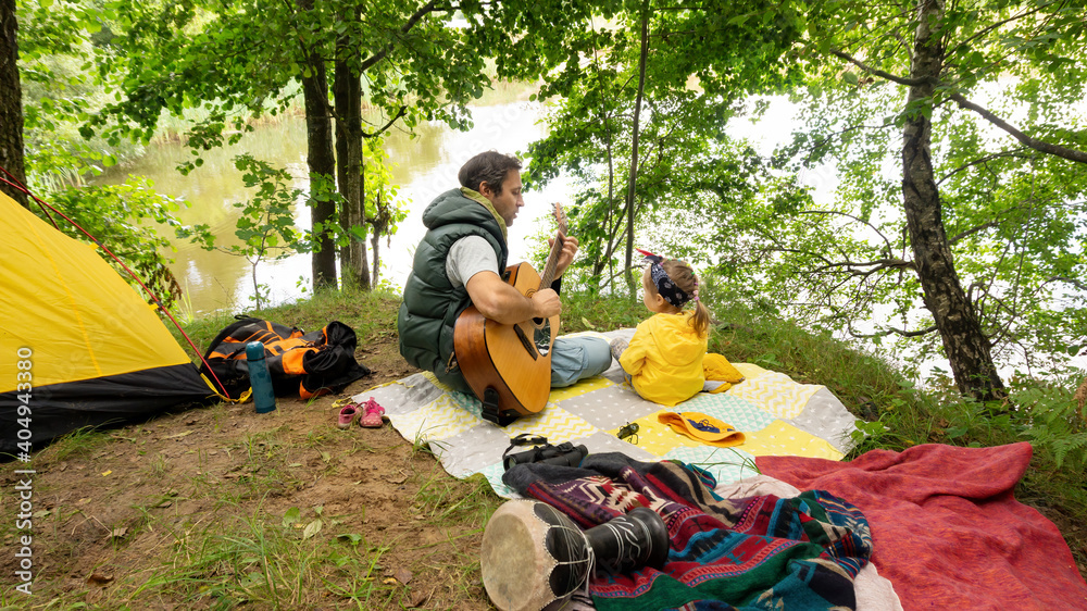A father plays the guitar for his little daughter while vacationing at the family camping on the shore of a lake. Rest during tourist trekking around the forest lake. Outdoor music lessons. A father plays the guitar for his little daughter while vacationing at the family camping on the shore of a lake. Rest during tourist trekking around the forest lake. Outdoor music lessons.