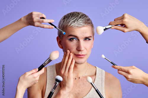 make-up of transgender male model isolated on purple background, man looks at camera touching face, in doubts