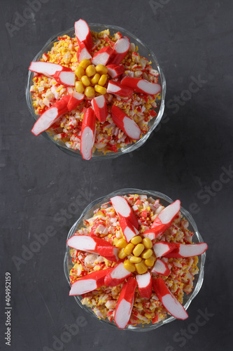 Two servings of salad with crab sticks, eggs and corn in transparent glass bowls on dark background. Closeup