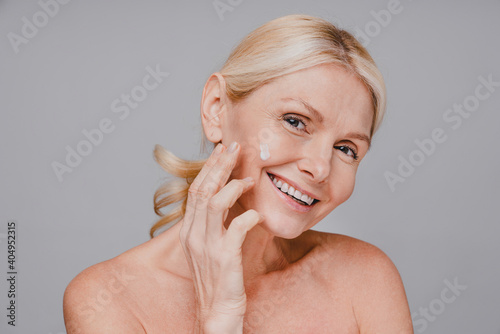 Smiling charming aged woman rubbing face cream isolated over grey background