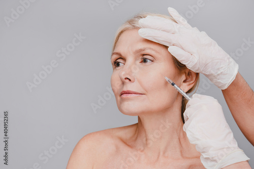 Good-looking mature caucasian woman undergoing facial beauty injections isolated over grey background photo