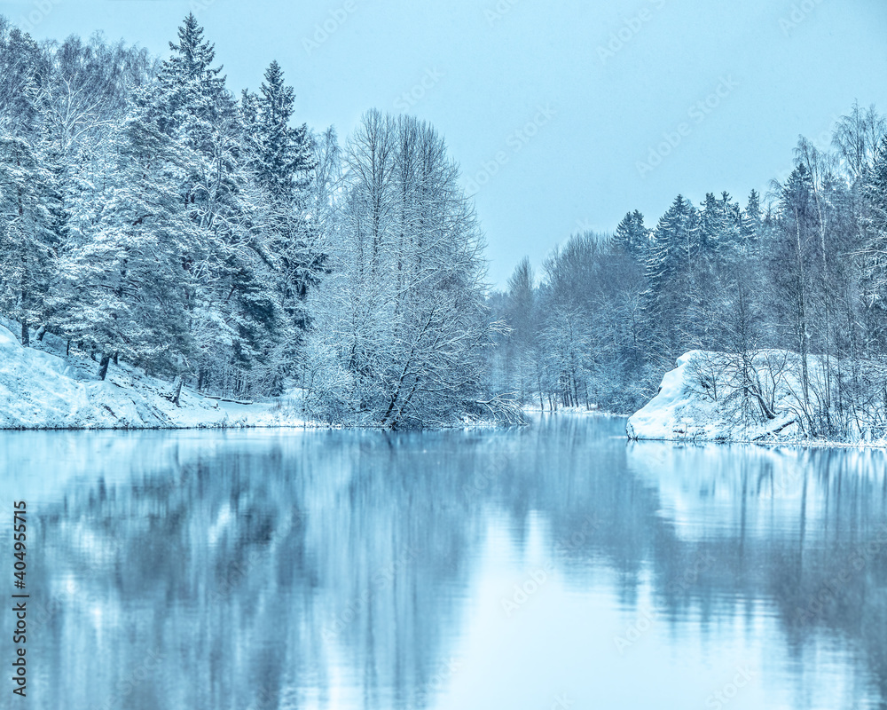 Beautiful tranquil winter scenery. Snowy river coastline. View on frozen lake coast. Trees on the shore covered with snow and reflected in lake. January scene. Frosty winter day. Blue toning. Snowfall