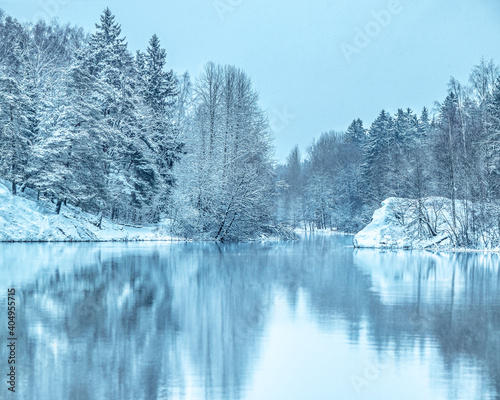 Beautiful tranquil winter scenery. Snowy river coastline. View on frozen lake coast. Trees on the shore covered with snow and reflected in lake. January scene. Frosty winter day. Blue toning. Snowfall