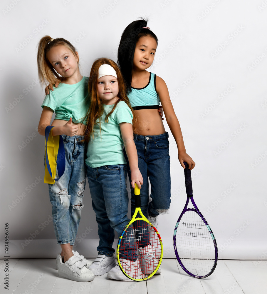 Happy sportive kids enjoy workout and healthy lifestyle. Little girls wearing jeans, sportive t-shirts holding water bottle, big tennis racket, fitness rubber showing thumbs-up. Isolated studio shot