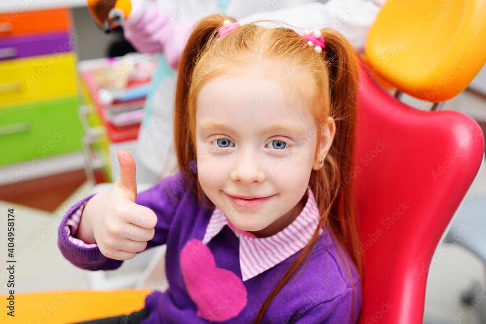 baby little red-haired girl sitting in a dental chair smiling and shows a thumb-sign class happy with the result of treatment.