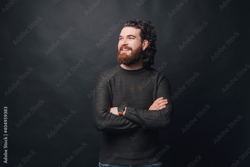 Handsome man with beard and long curly hair standing over dark background with crossed arms and looking away.