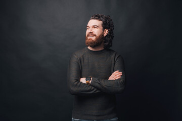 Handsome man with beard and long curly hair standing over dark background with crossed arms and looking away.