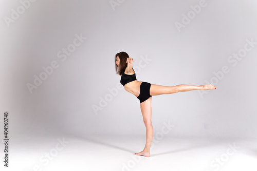 A slender girl in a black sports swimsuit on a white isolated background. Girl athlete stands on one leg on a white isolated background with place for text or logo. The dancing girl on a white backgro