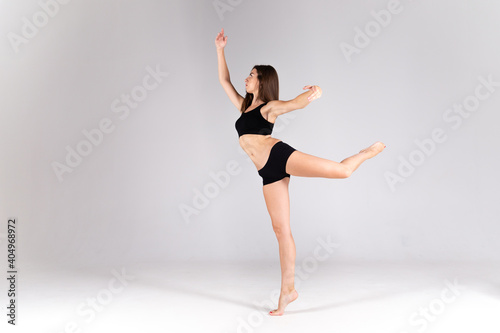 A slender girl in a black sports swimsuit on a white isolated background. Girl athlete stands on one leg on a white isolated background with place for text or logo. The dancing girl on a white backgro