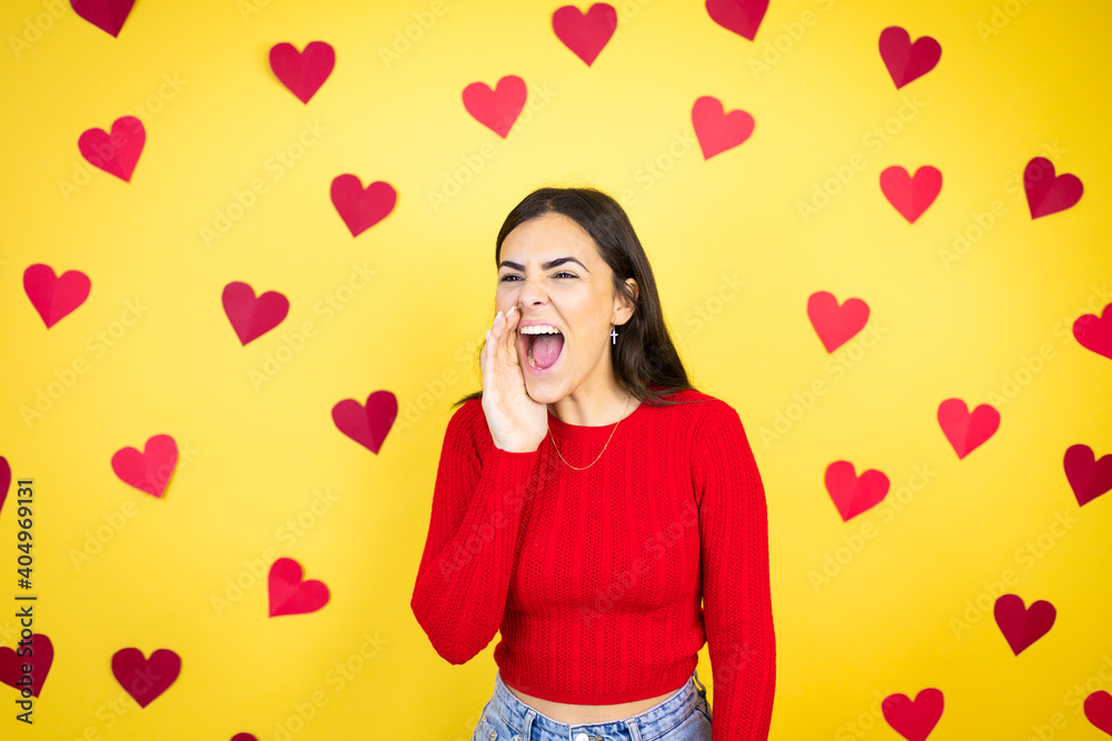 Young caucasian woman over yellow background with red hearts shouting and screaming loud to side with hand on mouth Young caucasian woman over yellow background with red hearts shouting and screaming loud to side with hand on mouth