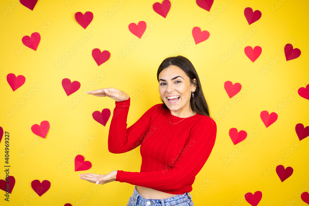 Young caucasian woman over yellow background with red hearts gesturing with hands showing big and large size sign, measure symbol. Smiling . Measuring concept. Young caucasian woman over yellow background with red hearts gesturing with hands showing big and large size sign, measure symbol. Smiling . Measuring concept.