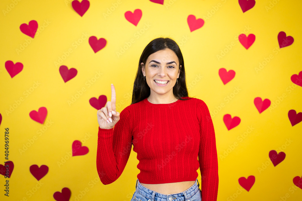 Young caucasian woman over yellow background with red hearts showing and pointing up with fingers number one while smiling confident and happy Young caucasian woman over yellow background with red hearts showing and pointing up with fingers number one while smiling confident and happy