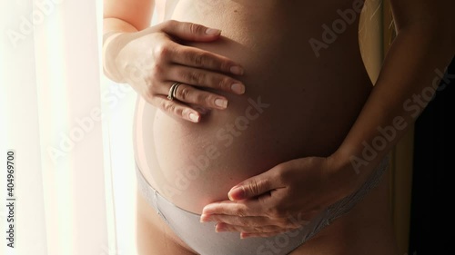 Closeup of pregnant woman gently touching and stroking her growing tummy next to big window in bedroom at home. Concept of happy pregnancy and baby anticipation