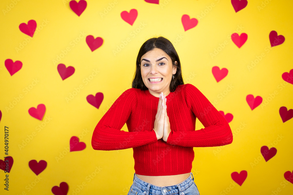 Young caucasian woman over yellow background with red hearts begging and praying with hands together with hope expression on face very emotional and worried Young caucasian woman over yellow background with red hearts begging and praying with hands together with hope expression on face very emotional and worried
