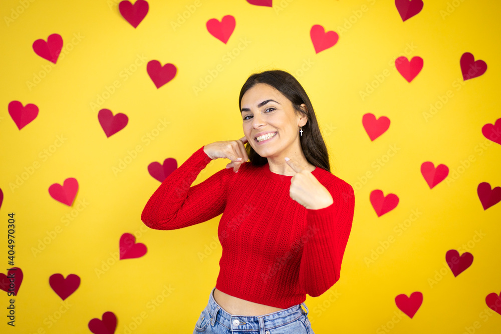 Young caucasian woman over yellow background with red hearts doing the “call me” gesture with her hands. Young caucasian woman over yellow background with red hearts doing the “call me” gesture with her hands.