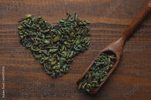 Dried green tea leaves in the shape of heart and spoon on a wooden background. Selective focus, top view, flat lay