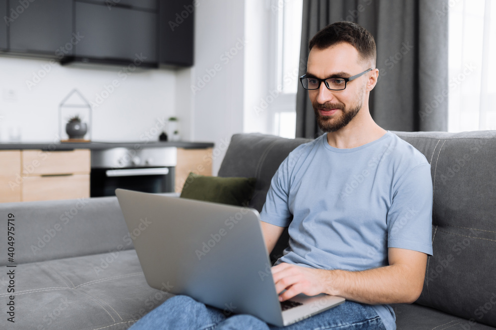 Confident male freelancer working on project from home, searching information in the network. Caucasian male manager or IT specialist sitting on the couch, using laptop, chatting with friends or Confident male freelancer working on project from home, searching information in the network. Caucasian male manager or IT specialist sitting on the couch, using laptop, chatting with friends or