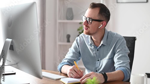 A young man using trendy PC for working, studying, watching online webinars. A confident entrepreneur in glasses looks at the screen sitting at the desk and taking a notes carefully