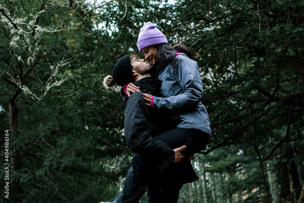 The young Caucasian woman in the pink hat kisses her boyfriend while he holds her in his arms. They are on a beautiful wooden path by a mountain trail.