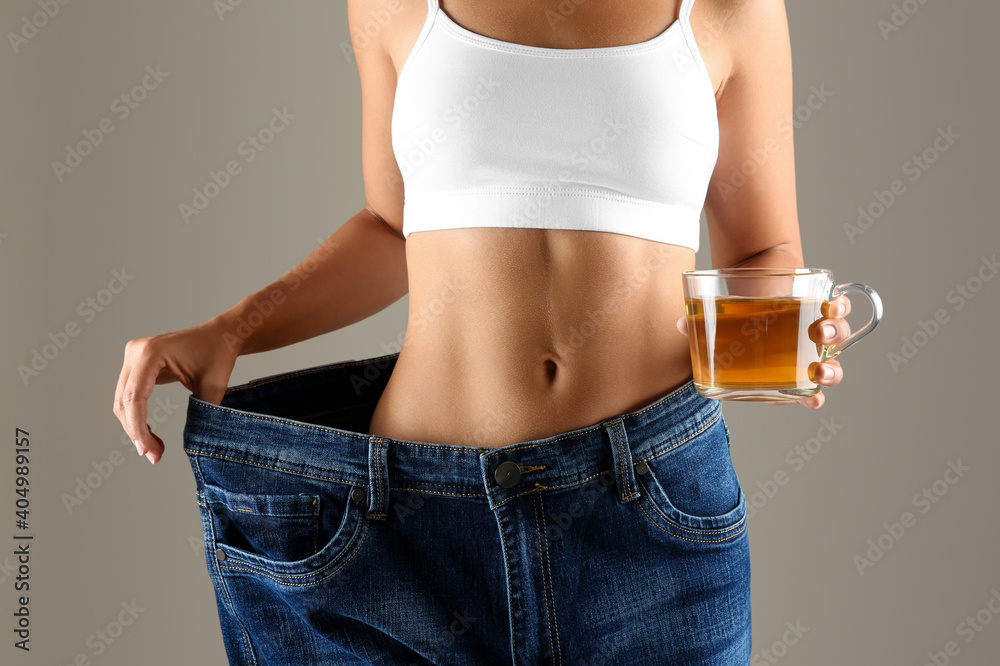 Young woman in old big jeans with cup of tea showing her diet results on beige background, closeup - obrazy, fototapety, plakaty 