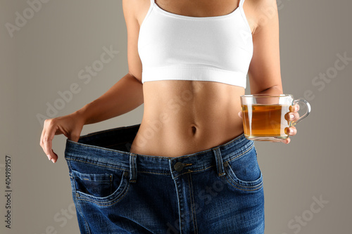 Young woman in old big jeans with cup of tea showing her diet results on beige background, closeup