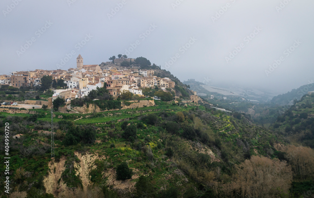 Foggy morning at the hill of Polop de Marina with church and castell over green forest, Spain