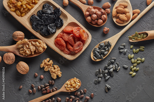 Different kinds of nuts, dried fruits in wooden spoones and dish on black slate background. Top view. Healthy food. Vegetarian nutrition