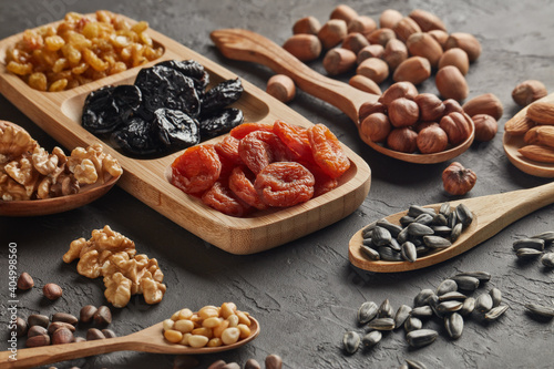 Different kinds of nuts, dried fruits in wooden spoones and dish on black slate background. Healthy food. Vegetarian nutrition