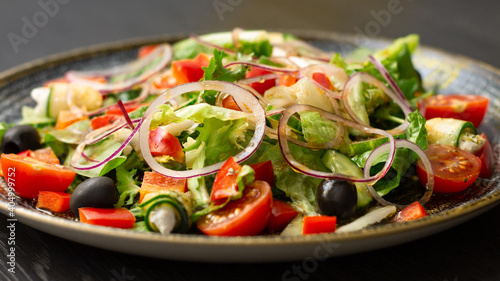Vegetable salad with greens and olives, in dark clay plate on wooden table