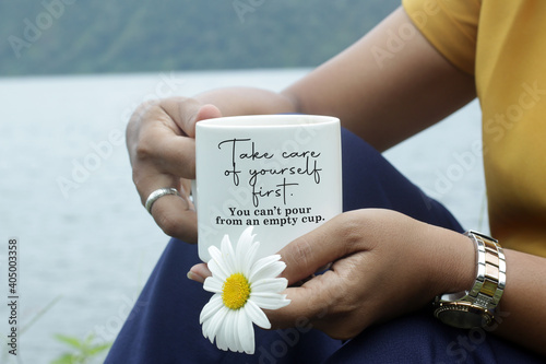 Inspirational quote - Take care of yourself first. You cannot pour from an empty cup. With young woman sitting in the lake holding cup of tea or coffee and a flower. Self love and care concept. photo