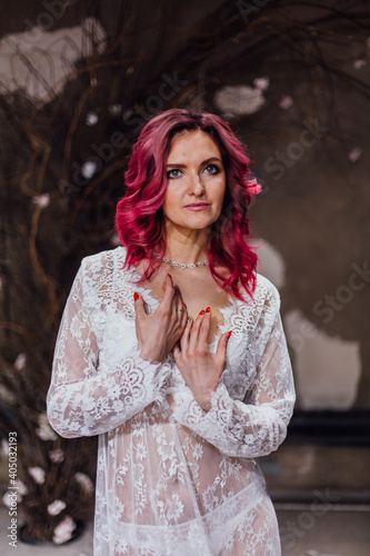 Attractive woman with pink hair in white light dress posing in a dark room with concrete walls. Lady in gentle lace peignoir