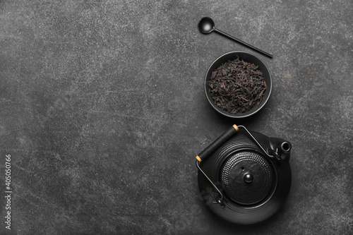 Dry black tea leaves in bowl, teapot and spoon on dark background