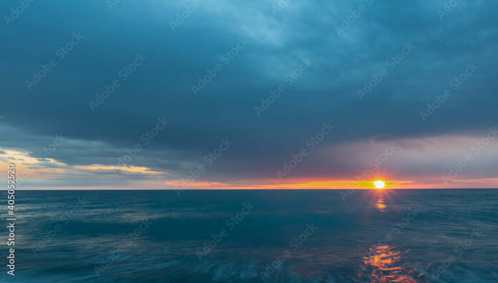 Dense blue clouds and turquoise sea. Between the sky and the water, the setting sun illuminates the sky orange. Sunny path. Black Sea