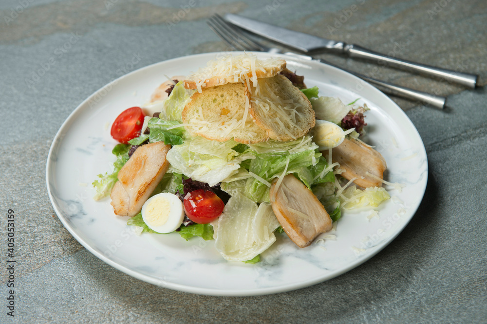 Close up of Traditional Italian Caesar salad served with fork, knife on marble plate. Salad with bread croutons, grilled chicken fillet slices, fresh lettuce, quail eggs, tomatoes on stone background
