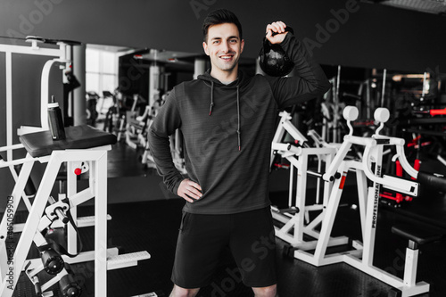 Photo of a young coach who looks at the camera with a smile and lifts a weight with ease while standing in the middle of the gym