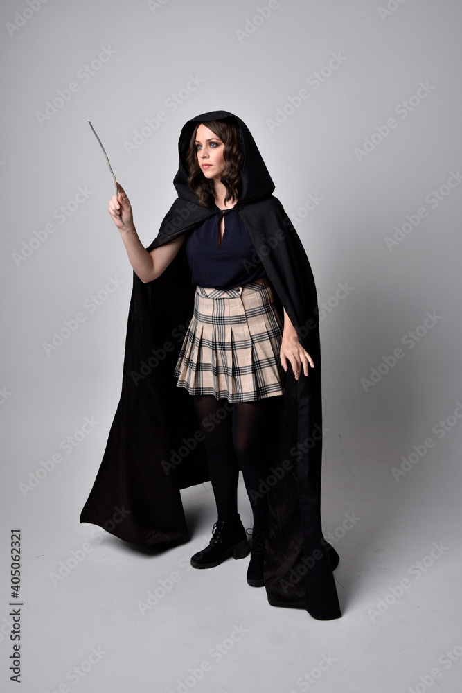 Naklejka premium full length portrait of pretty brunette woman wearing tartan skirt and boots with long black cloak.. Standing pose holding a magical wand against a studio background.