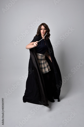 full length portrait of pretty brunette woman wearing tartan skirt and boots with long black cloak.. Standing pose holding a magical wand against a  studio background.