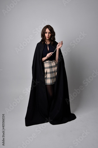 full length portrait of pretty brunette woman wearing tartan skirt and boots with long black cloak.. Standing pose holding a magical wand against a studio background.