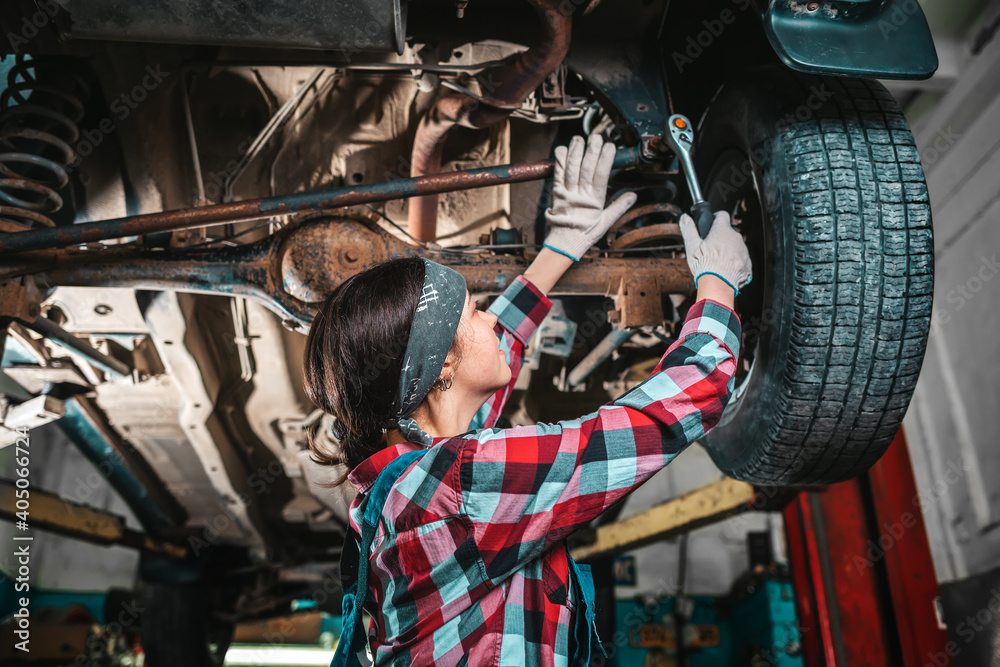 Portrait of a young female mechanic in uniform and gloves who repairs a car. The car is on the lift. Bottom view