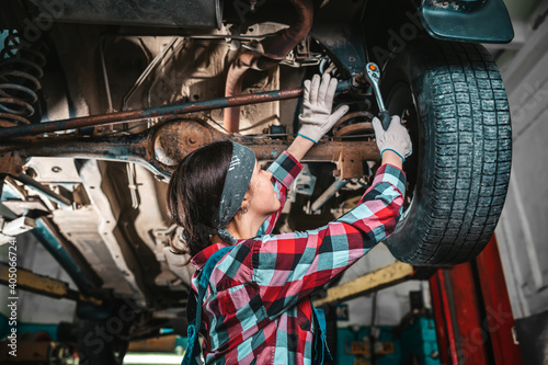 Portrait of a young female mechanic in uniform and gloves who repairs a car. The car is on the lift. Bottom view