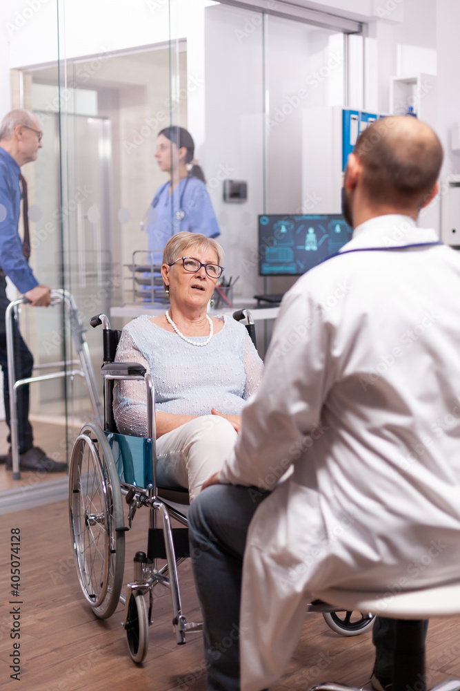 Paralyzed discussing with geriatrician sitting in wheelchair in the course of examination. Medical specialist wearing stethoscope discussing with invalid elderly woman in consultation room of private.