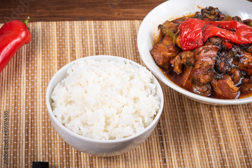 Spicy stewed pork with mushrooms in oriental style with spices, hot pepper in a bowl and a cup of boiled rice on a bamboo napkin and chopsticks - top view, close-up photo