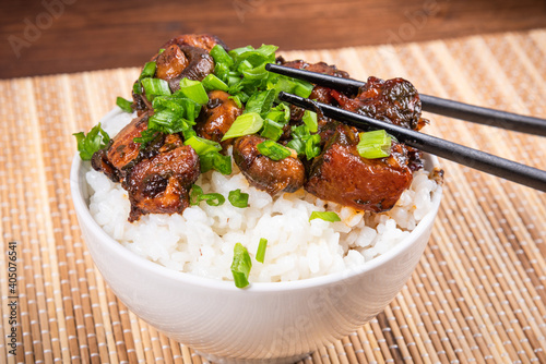 Small bowl with spicy stewed pork with mushrooms in oriental style with spices, hot peppers and boiled rice sprinkled with chopped green onions on a bamboo napkin and chopsticks - top view, close-up