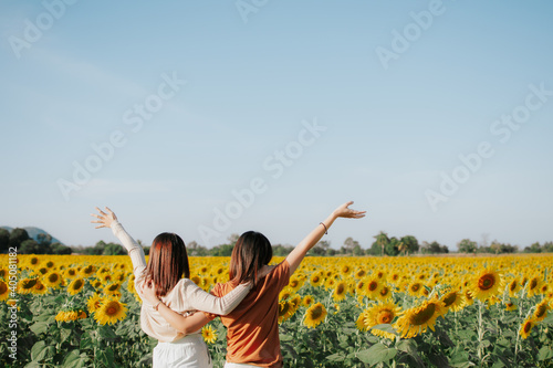 Two women are standing in a field of sunflower and hugging each other. Their hands are raised up to the sky. Friendship concept and copy space.