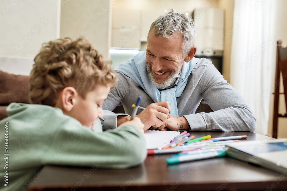 Happy grandfather and grandson sitting at table, having fun while drawing with colored felt tip pens together at home Happy grandfather and grandson sitting at table, having fun while drawing with colored felt tip pens together at home