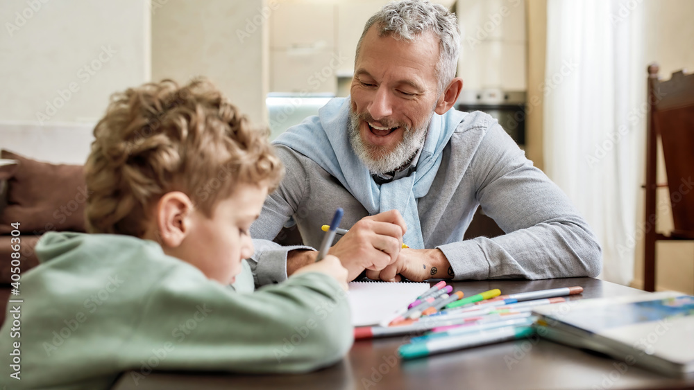 Happy grandfather enjoying spending time with his little grandson, drawing with colored felt tip pens on paper while sitting at table - obrazy, fototapety, plakaty 