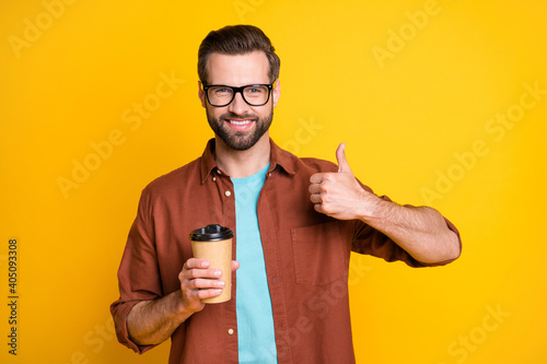 Photo portrait of bearded millennial in eyewear showing like sign keeping beverage cup takeaway isolated on bright yellow color background