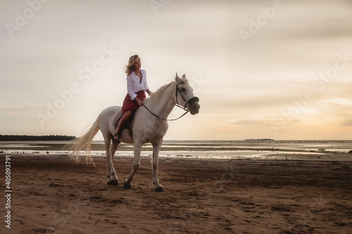 Cute happy young woman on horseback in summer beach by sea. Rider female drives her horse in nature on evening sunset light background. Concept of outdoor riding, sports and recreation. Copy space